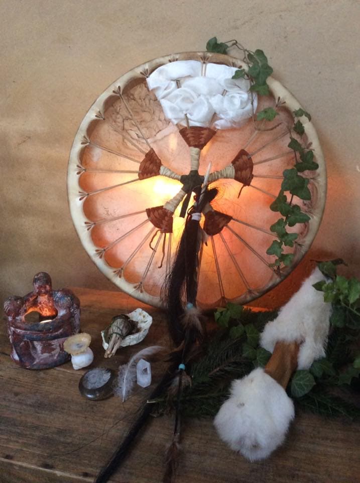 Tambour chamanique orné de fleurs blanches et de lierre, posé parmi bougie, pierres, plumes et fourrure
