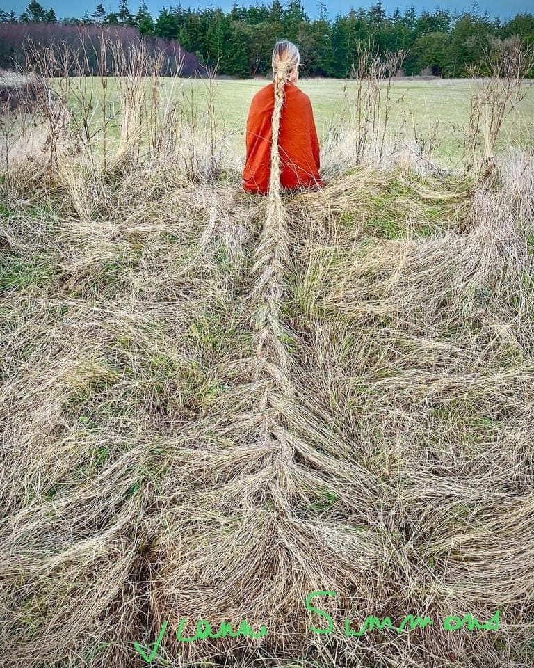 Land art : une personne en orange assise dans un champ, sa longue tresse se prolongeant en herbes tressées