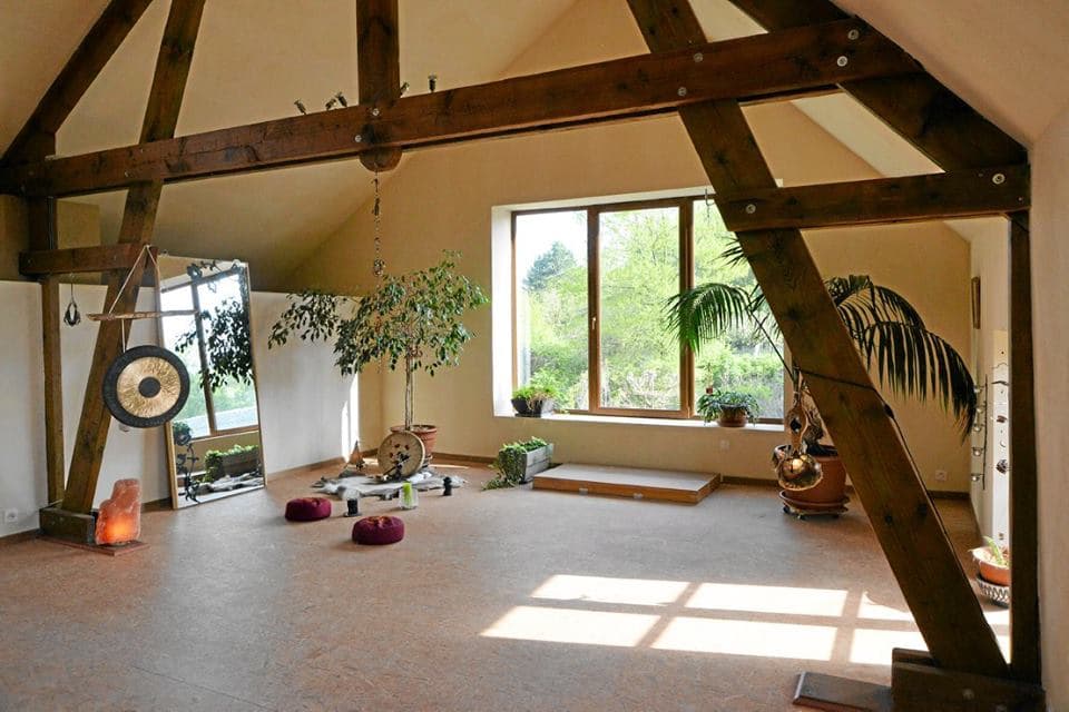La salle de pratique vue depuis l’entrée : charpente apparente, gong, plantes et baie vitrée sur le jardin