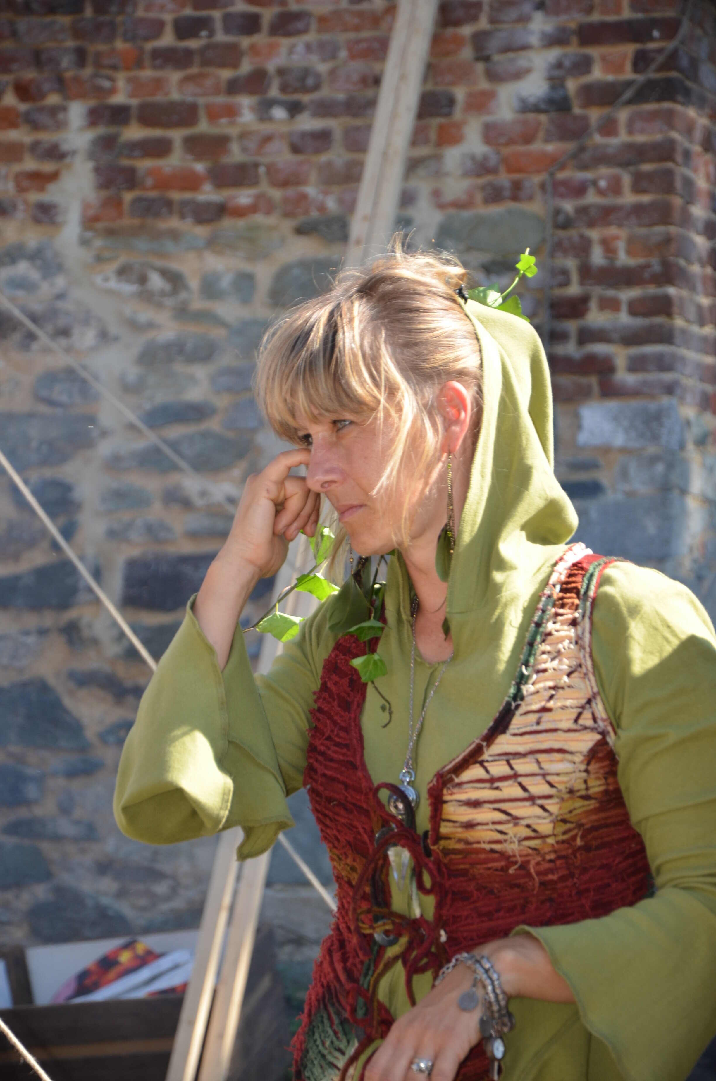 Véronique Linclau de profil, pensive, en tunique verte à capuche et gilet brodé, devant un mur de briques