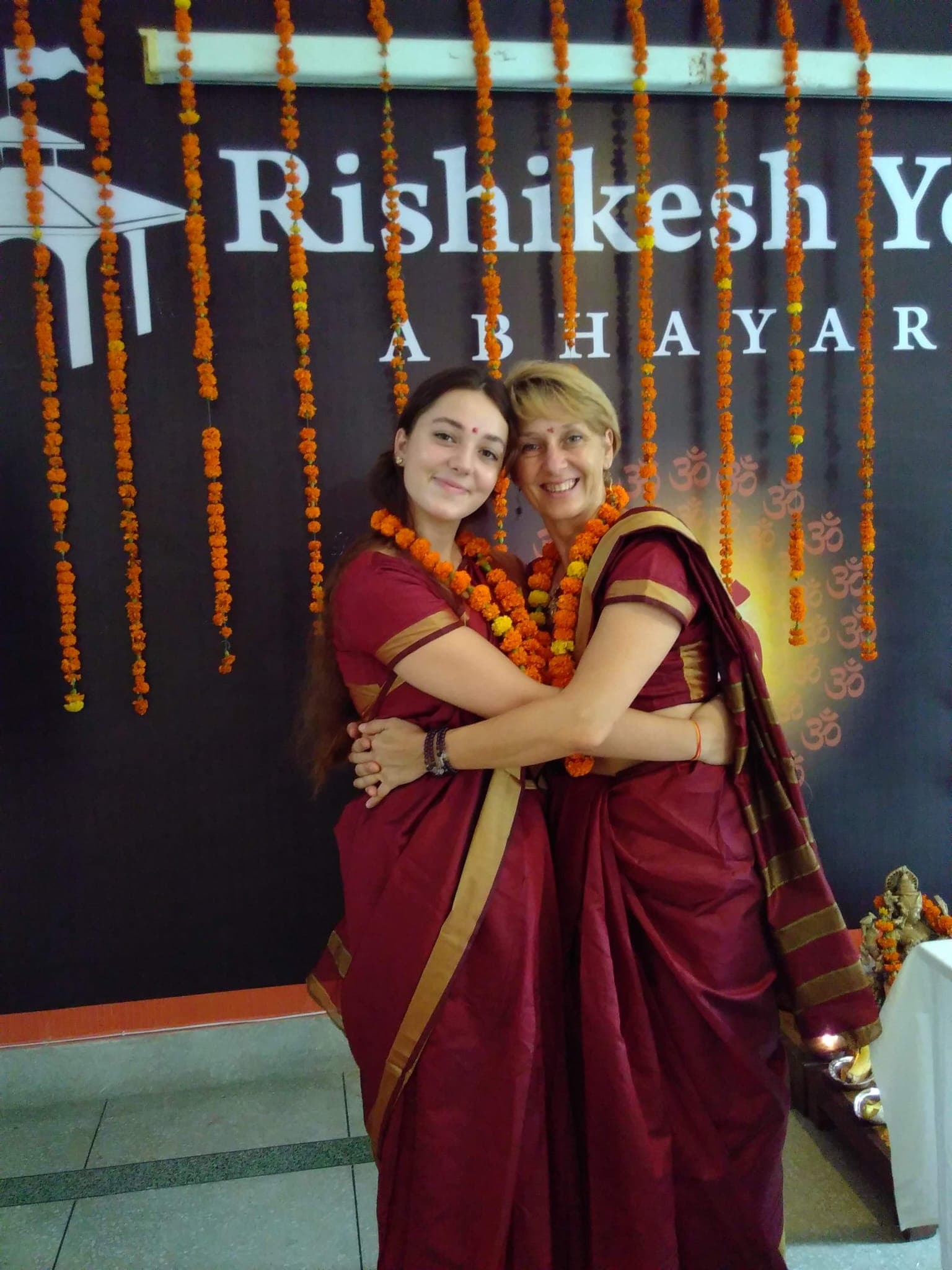 Nell et Véronique enlacées, saris bordeaux et guirlandes orange, devant l’enseigne de l’école Rishikesh Yogpeeth
